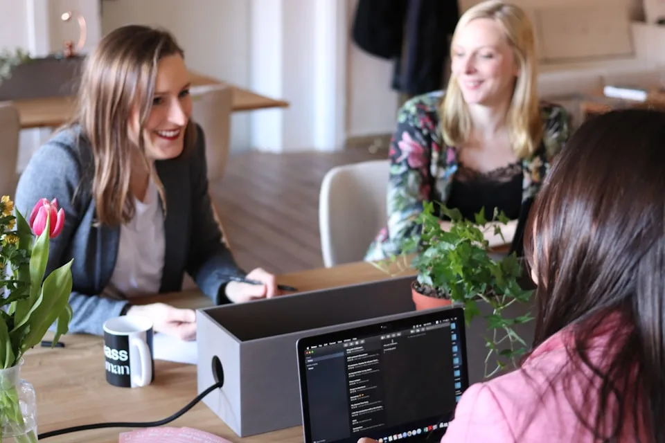 Três mulheres conversam ao redor de uma mesa com computador e materiais de trabalho.