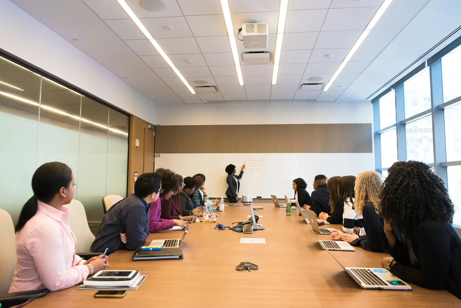 Grupo de profissionais acompanha uma explicação em um quadro branco, ao redor de uma mesa de reunião.