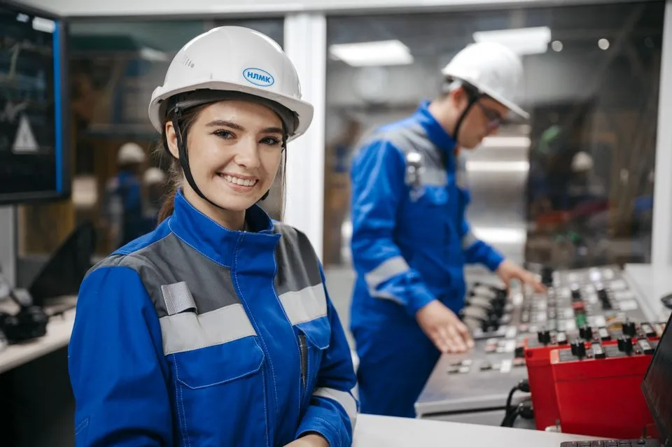 Profissional sorrindo em primeiro plano, com uniforme e capacete de jugular afivelada, diante de colegas em uma sala de controle industrial