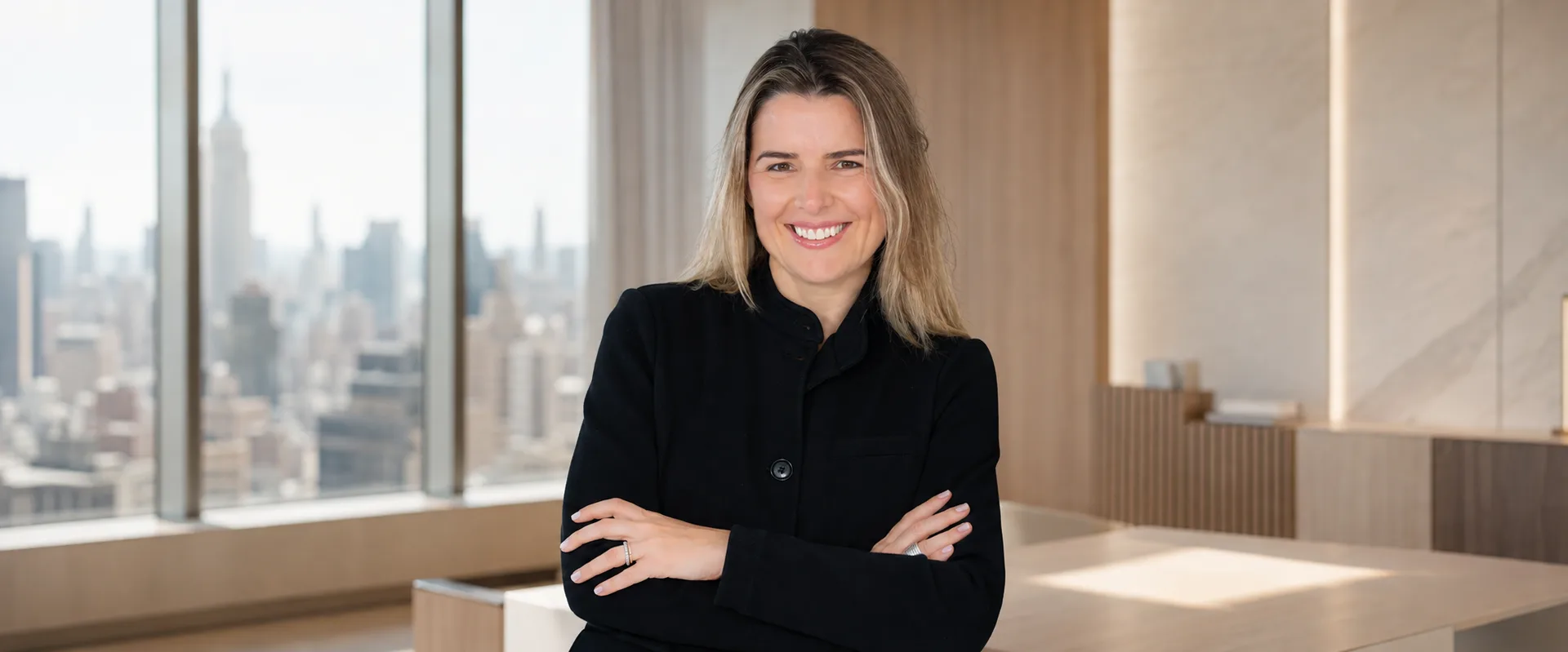 Andreza Araújo sorrindo, de blazer preto e braços cruzados, em composição de escritório em Nova York
