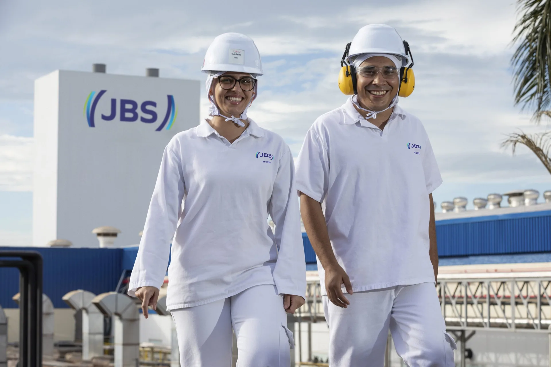 Dois colaboradores da JBS com uniformes e equipamentos de proteção, em frente a uma instalação da empresa