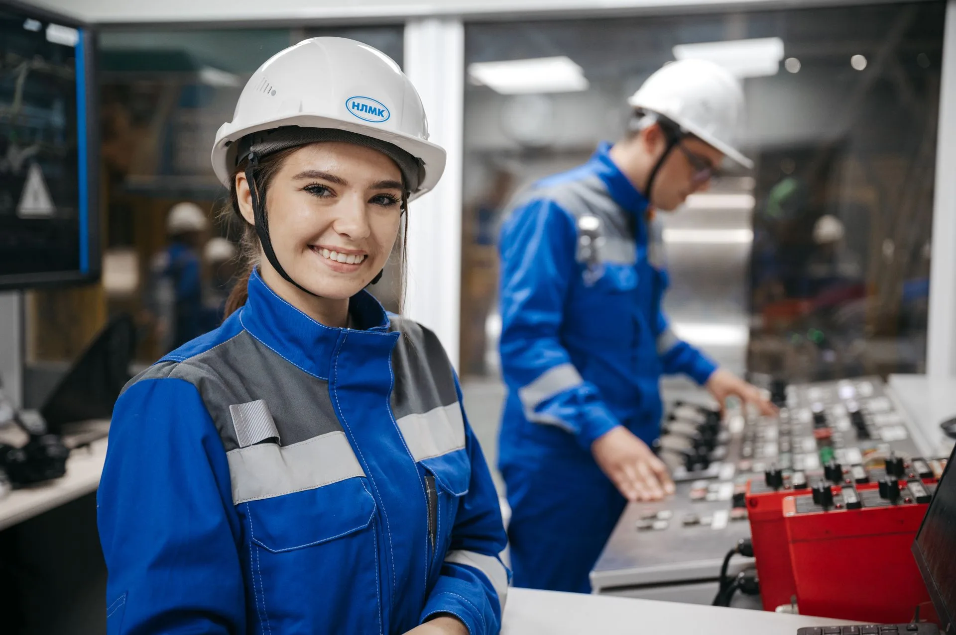 Profissional sorrindo em primeiro plano, com uniforme e capacete de jugular afivelada, diante de colegas em uma sala de controle industrial