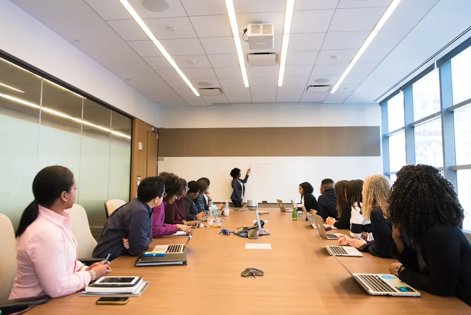 Grupo de profissionais acompanha uma explicação em um quadro branco, ao redor de uma mesa de reunião.