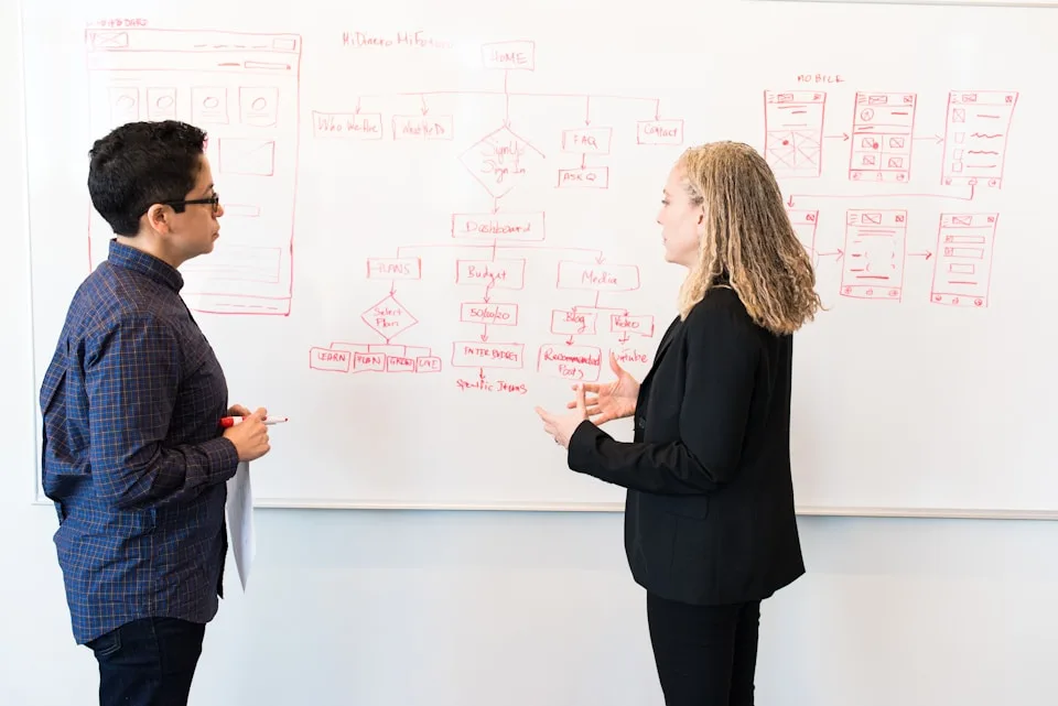 Dois profissionais conversam diante de um quadro branco com diagramas e anotações.